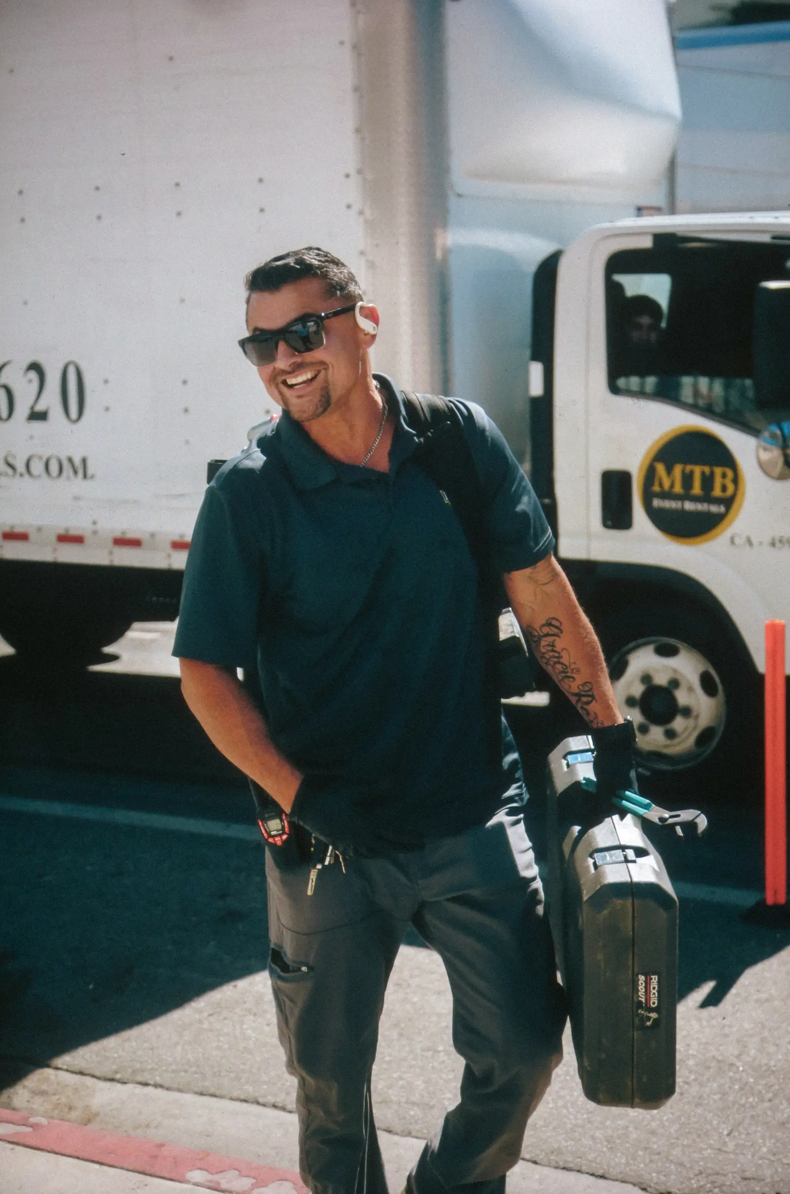 A smiling plumber carries a RIDGID tool case past a work truck.